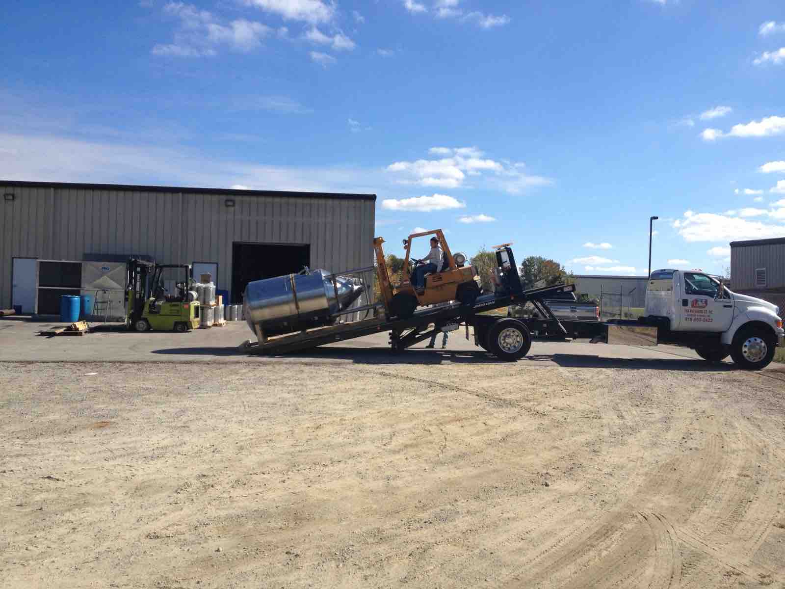 Steel tank being loaded onto a rollback truck beside a forklift